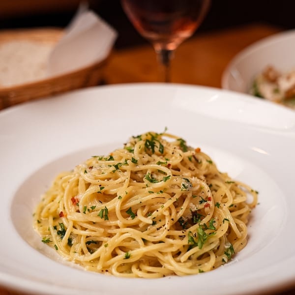 Spaghetti pasta with parsley and cheese in a white bowl, with bread and wine in the background.