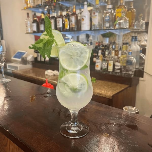 A refreshing mojito with lime slices and mint sits on a wooden bar counter with liquor bottles in the background.