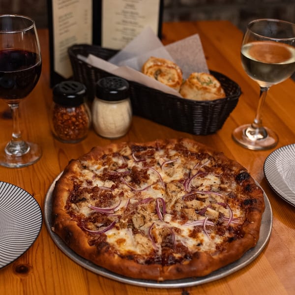 A whole pizza with red onions and chicken, two glasses of wine, and bread on a wooden table.