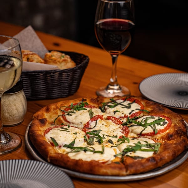 A Margherita pizza with fresh basil and tomatoes, alongside wine glasses and bread on a wooden table.
