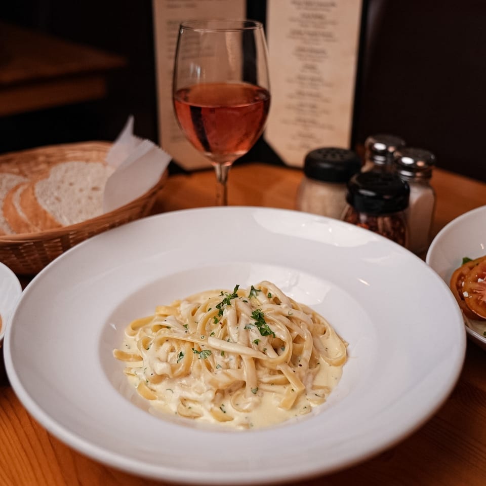 Creamy fettuccine alfredo with parsley, a glass of rosé, bread, and spices on a wooden table.