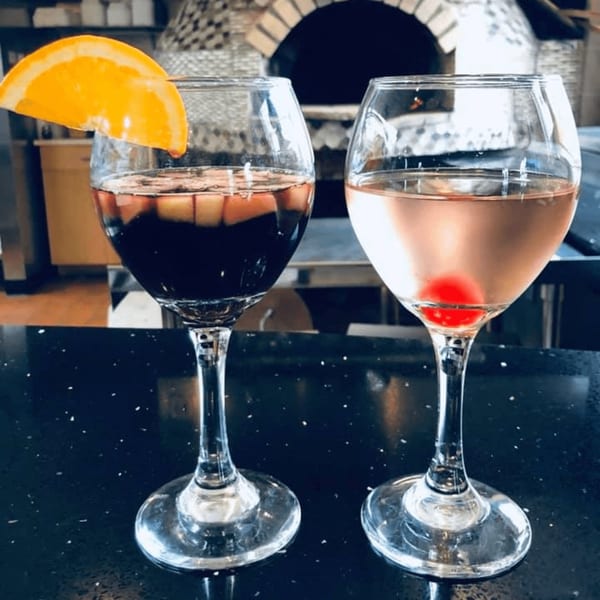 Two cocktails, a dark sangria and a pink drink with a cherry, in front of a pizza oven.